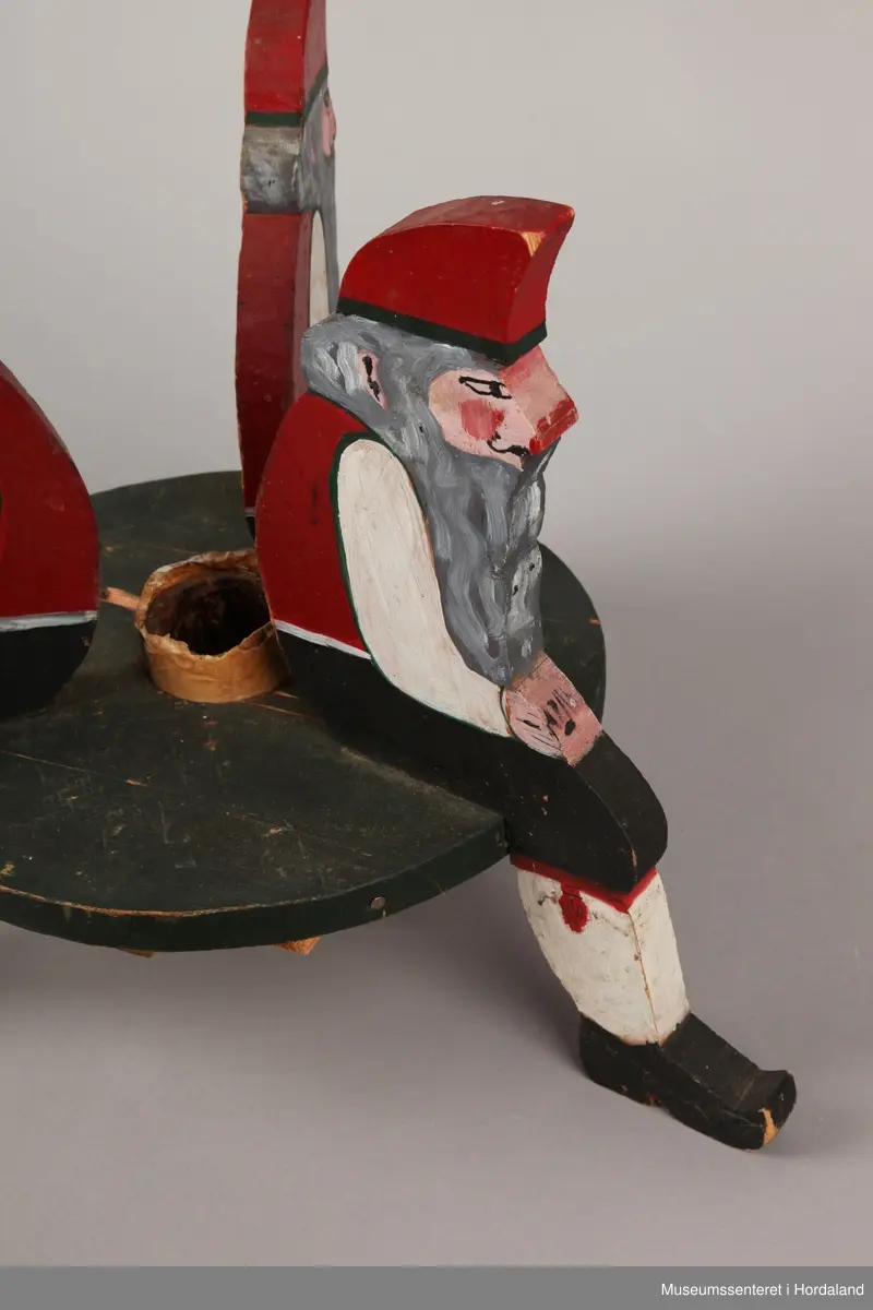 3 nisser i utskårne i tre som sit på ei rund plate. Nissaane og plata er skårne ut i 2 cm tjukk plank. Nissane er malte i ulike fargar. Plata er malt svart. Brun tape rund holet til juletreet.