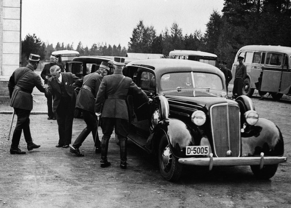 Elverum, Kong Haakon 7 og kronprins Olav i militæruniformer går inn i bilen til drosjeeier Vold, Chevrolet med registreringsnummer D-5005 årsmodell 1935 ved skolen i Strandbygdveien. Kronprins Olav hilser på drosjeeier Vold. General Sande til høyre.