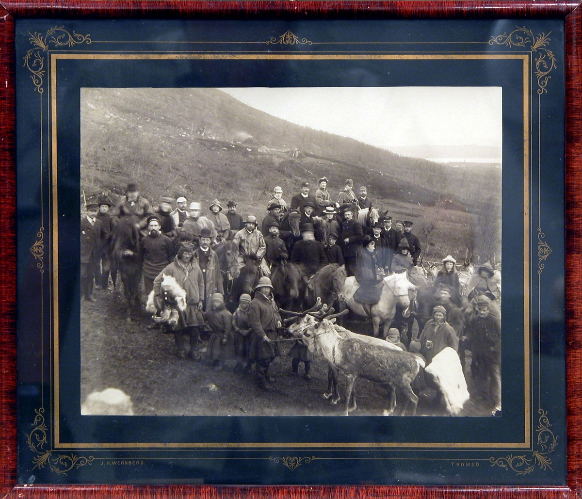 Gruppebilde av reisende og besetning fra D/S "Sigurd Jarl" tatt ute i bart landskap, sammen med noen fra den lokale samiske befolkning med hester og reinsdyr.