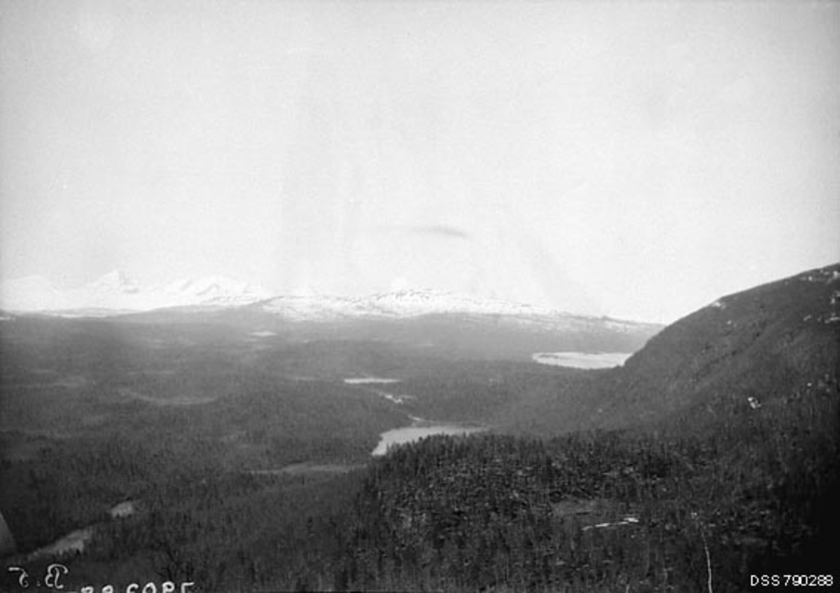 Landskapsfotografi, utsiktsbilde fra Nordgårdåsen i Målselv kommune i Troms. Opptaket antas å være gjort omkring 1910. Fotografen har vendt kameraet vestover. Vi ser noe av vannspeilet på Lille Rostavatn litt til høyre for bildets midtpunkt og skimter de på dette tidspunktet snødekte fjellene på den nordøstre sida av dette vatnet i bakgrunnen. I dalbotnen, nærmere fotografen aner vi et vassdrag.