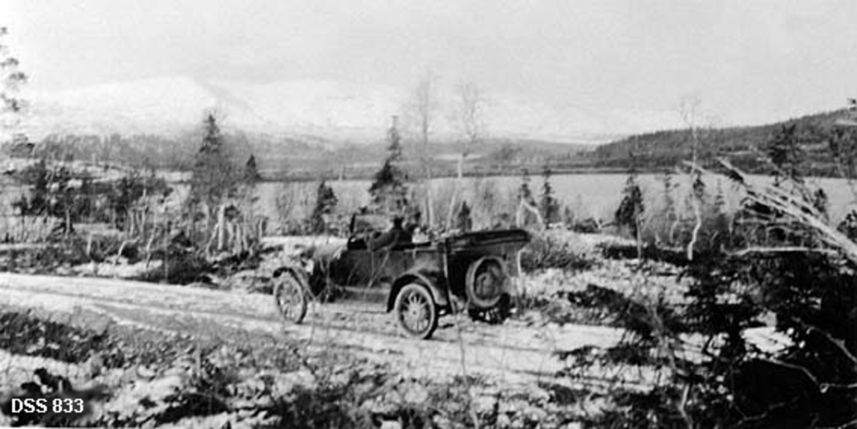 Utsikt fra fjellovergang mellom Fiplingdal og Svenningdal på Sør-Helgeland.  I forgrunnen en veg, der det står en personbil med nedtrukket kalesjetak og sjåfør i førersetet.  Landskapet i forgrunnen er lyngbevokst med enkelte bjørke- og bartrær.  Bakenfor skimtes ei sjøflate.  Bakenfor varnet et skogbelte, og helt i bakgrunnen snødekte snaufjell.