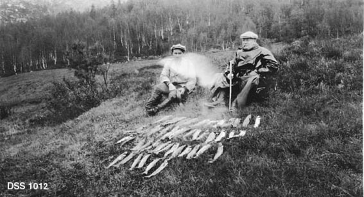 Skogforvalter Petter Magnus Vikhammer (1883-1952) og skoginspektør Einar Emil Nilsen (f. 1865) sittende på bakkekam med mye fin fisk etter ei natts garndrett i Hundålvatnet.  Vikhammer er iført vindjakke, Nilsen har svart regnfrakk og spaserstokk.  De sitter på et oppdyrket engareal.  I bakgrunnen en skråning med bjørkeskog og enkelte grantrær. 