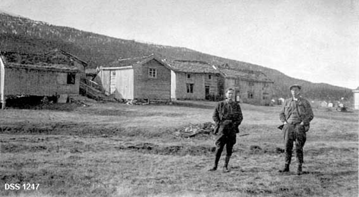 Gardsanlegget på Øvre Svenningdal i Grane på Helgeland.  Fotografiet er tatt fra ei grasslette hvor det sto to menn med et gardstun i bakgrunnen.  Tunet omfattet (fra venstre) to løer (den ene delvis skjult) en loftsbygning og to våningshus som var bygd gavl mot gavl.  I bakgrunnen skimtes flere hus fra andre gardstun, og en skogbevokst åsrygg.