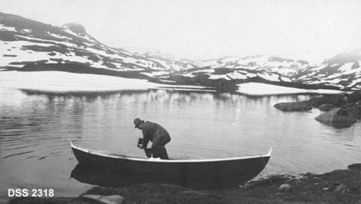 Parti fra Haukelisæter.  Mann som øser robåt ved bredden av et fjellvatn i forgrunnen, snaufjell med strimer av snø i bakgrunnen. 