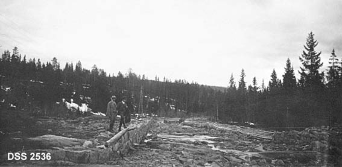 Ledeskjermer (skådammer) av stein (til venstre) og tre (til høyre) i vasstomt elvefar i Siljanvassdraget.  To menn står på steinskjermen.  Granskog i bakgrunnen.  Tidlig våropptak. 