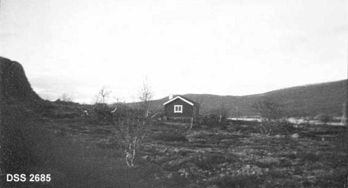 Blæstruds hytte nær Vålåsjøkampen på Dovrefjell.  Enetasjes fritidsbolig i viddelandskap.  Fjellskrent til venstre, sjø til høgre bak hytta. 