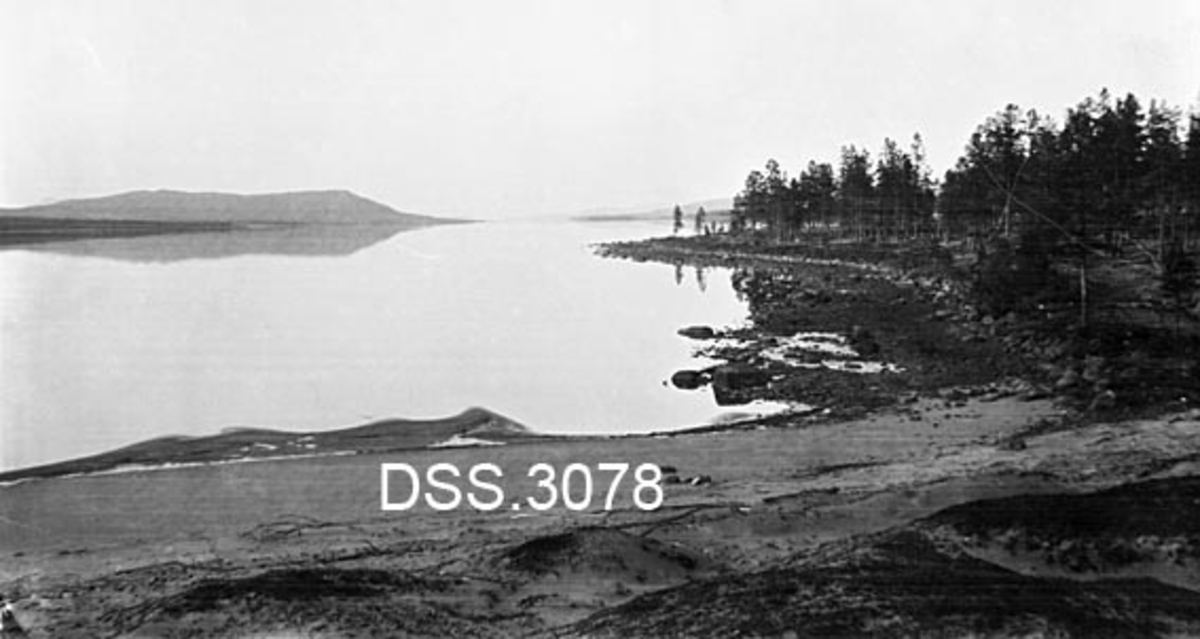 Utsikt nordover innsjøen Femund fra Femundsenden i Engerdal.  Lav vannstand.  Sandstrand i forgrunnen, mer steinrik strand oppover på østsida av sjøen.  Lav furuskog på land.  Bjørnberga (948 m. o. h. ) i bakgrunnen til venstre. 