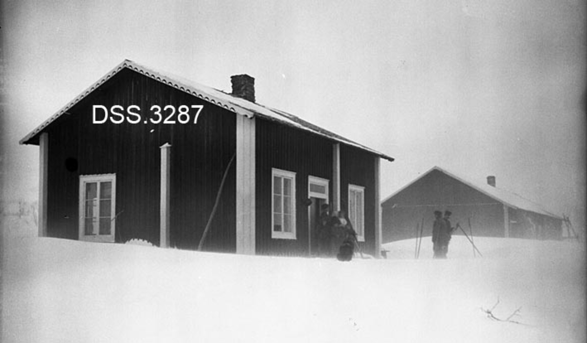 Merkenes fjellstue.  Vinterbilde, tatt i snøkav.  To bygninger, hvorav den nærmeste er et panelt laftehus, der laftekasser og dør- og vindusomramminger er kvitmalte.  Skiløpere foran inngangen.  Det andre huset trer ikke klart fram i bildet. 