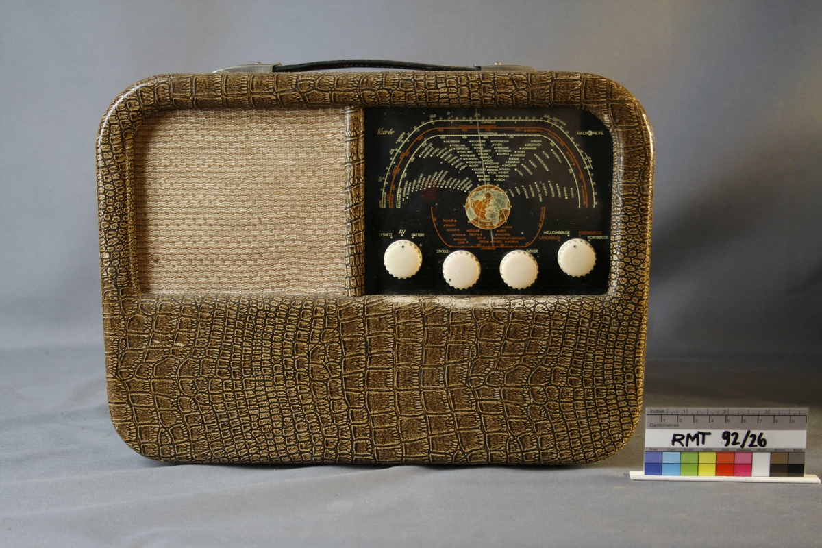 Rektangulær krokodillemønstret reiseradio med runde hjørner. Forside: skala med fire runde funksjons-, og styringsknapper. Høyttaler med trekk av tekstil. Håndtak på oversiden av radioen. Bakside: mangler bakplate. 6 uttak. I tillegg uttak for strøm. Bananstikk til antenne- eller platespilleruttaket ligger vedlagt, innpakket i plast.