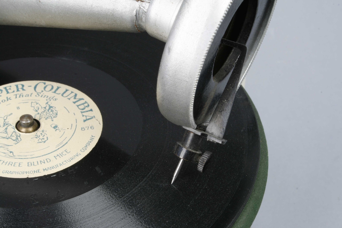 Leketøysgrammofon for avspilling av plater. Fjærmekanikk innvendig som trekkes opp for avspilling.
Nål og trakt som høytaler. 
Alle deler er i metall. Topplaten er malt grønn og limt rett på metall. Muligens en slags vinylmaling.