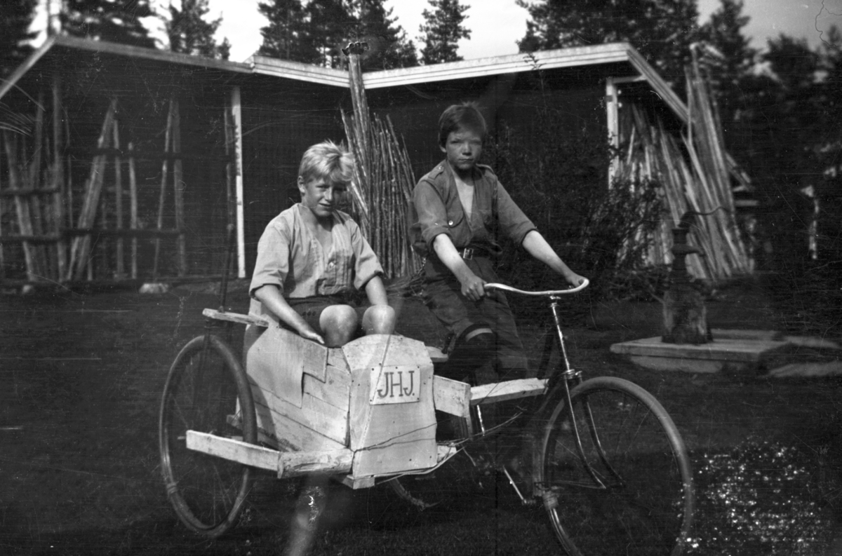 To gutter med sykkel og hjemmelaget sidevogn med skilt "JHJ". 
Vannpost bak.
Gutten til høyre er Jon Jørgensen (1908-1984), bror til Helge. Skiltet på sykkelen, "JHJ" henspeiler trolig på guttenes navn.
Gutten i "sidevogna" er Helge Jørgensen (1907-2004) fra Søbakken i Elverum. Identifiseringa er bekreftet av Helge Jørgensens sønn Anders Jørgensen.