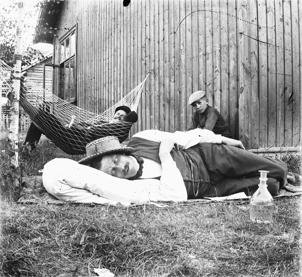 To menn og en gutt slapper av ved et hus. Den ene i ei hengekøye. En karaffel til høyre. 