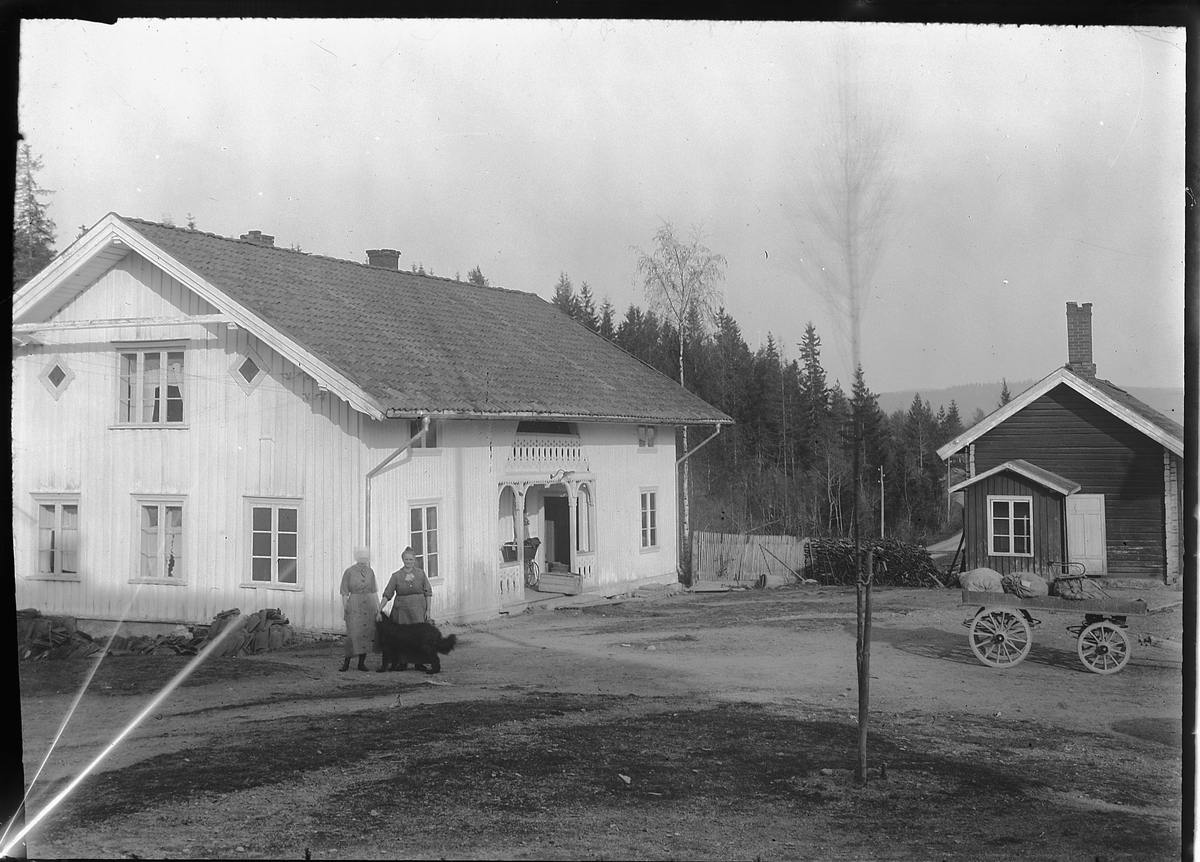 To kvinner med en hund på en gårdsplass. Ei hestekjerre til høyre. 