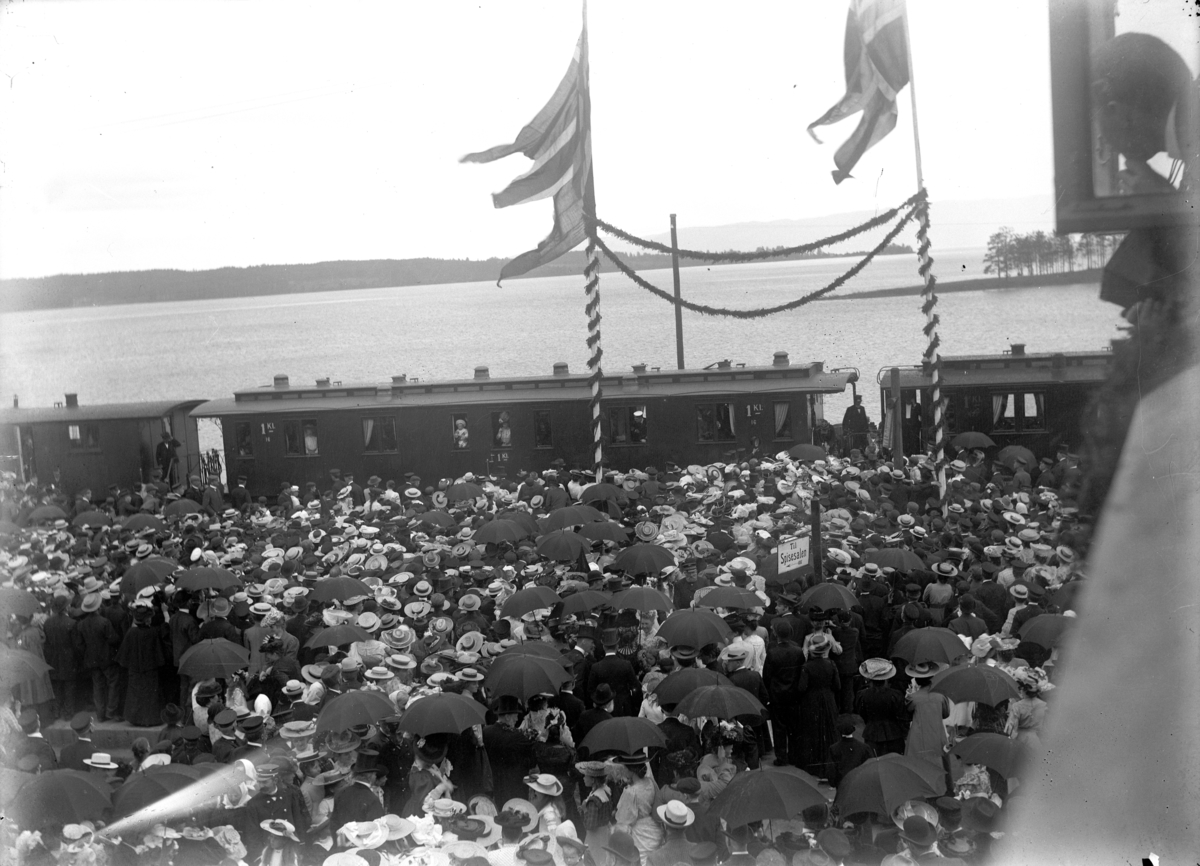 Hamar stasjon, kroningsreisen 1906, stor menneskemengde på perrongen, kong Haakon og dronning Maud ønskes velkommen, kongefamilien ombord i en jernbanevogn, girlandere og flagg