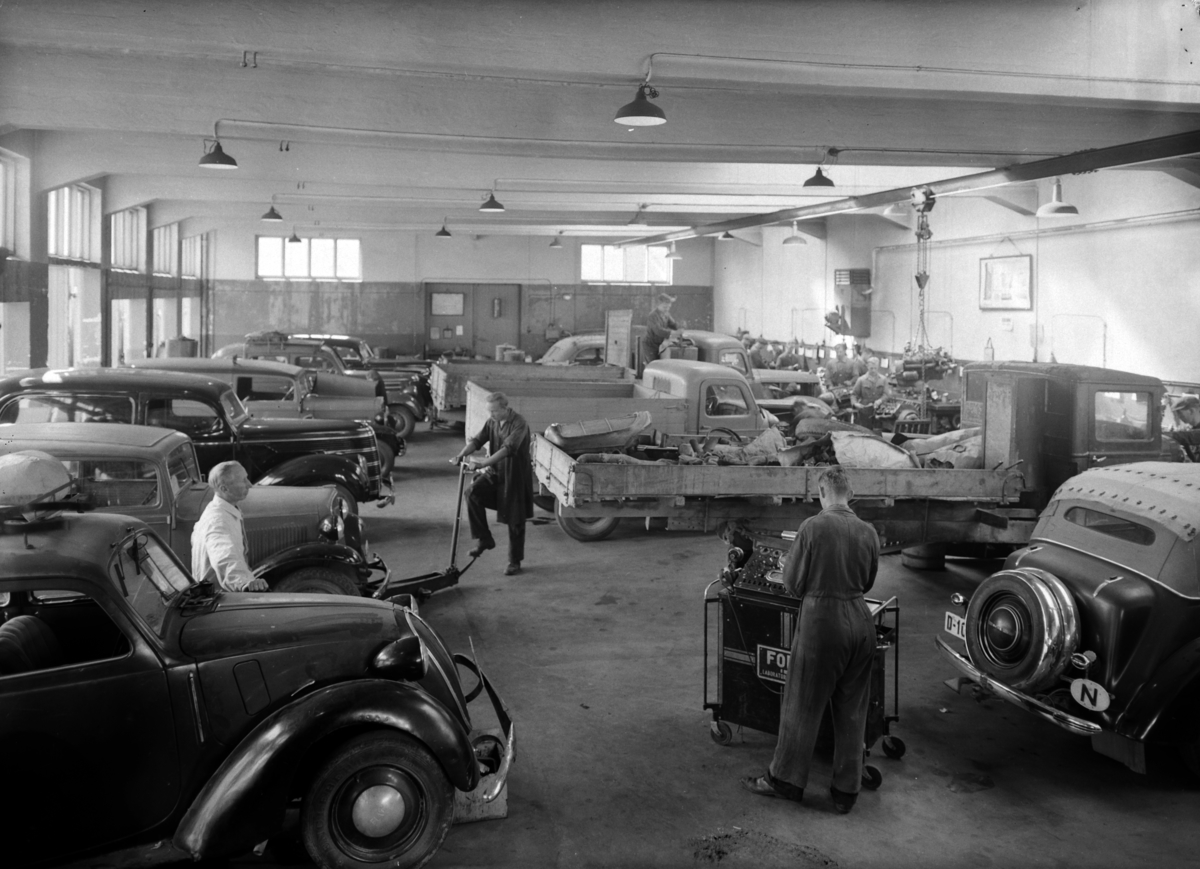 Hamar, Enggata 34, Oplandske Automobilforretning, Fordforhandler på Hamar siden 1923, bilmekanikkere i verkstedshallen, bilene fra venstre side, nærmest Fiat 1100 ca. 1936-39 modell, deretter ukjent, Ford V8 standard varebil (sedan). Til høyre bakparten på en Opel Super Six ca. 1936-38 modell,