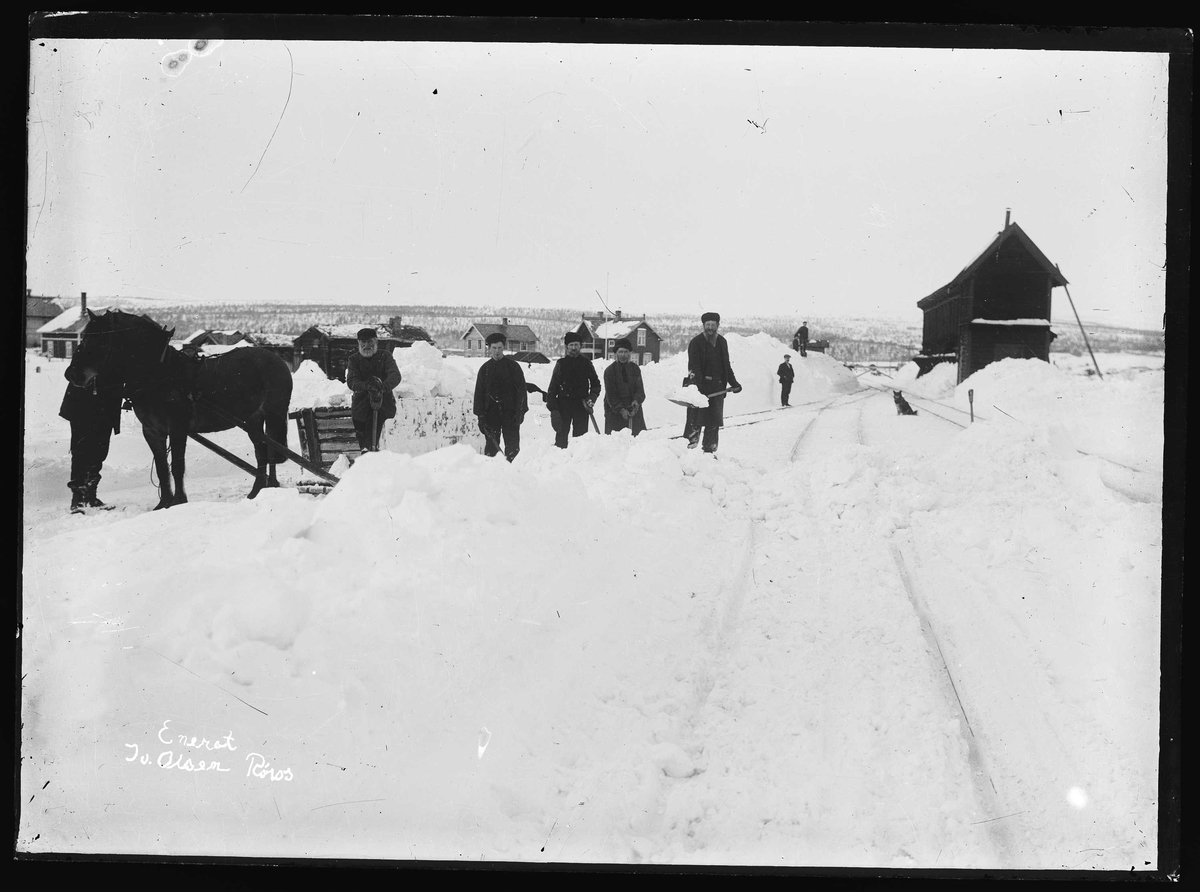 Snørydding ved Røros jernbanestasjon. Jernbanesporet ryddes for snø ved hjelp av spader og snøen kjøres bort med hest og slede, vest for jernbanestasjonen på Røros. Tømmerbygning som sees bak sleden er hovedbygninga til Eggagården/Kirkhusrommet. Uthuset til venstre. Bygningene står i dag mellom Slegghaugan og Falunveien. Den mørke bygningen midt i bildet bak arbeiderne ble i 1920 kjøpt av NSB og brukt som banemesterbolig.