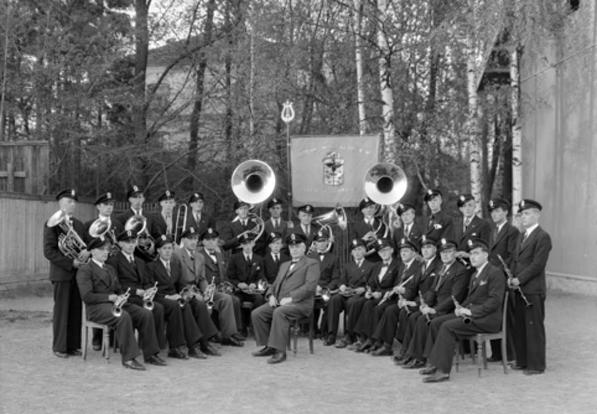 Hamar Musikkforening, stiftet 27. 10. 1921. Fane. Instrumenter. 
1. rekke f. v: Øivind Stømer, Tormod Nilsen, Kåre Rymo, Harald Jensen, Kristian Kristiansen, Bjørn Monsen, Arne Staff, dirigent Einar Schøyen, Georg Stenberg, Terje Søgaard, Hans Schøyen, Henry Nilsen, Harald Storm Myhr, Einar Bjærke, Egil Alex. Hansen. 
2. rekke f. v: Helge Aspeli, Jan Odvar Johnsen, Svein Staff, Arne Ransted, Sverre Engebretsen, Einar Bekkelund, Arthur Martinsen, Martin Olsen, Erik Larsen, Thor Monsen, Rolf Bakke, Henry Gulbrandsen, Arne Østbye, Tholeif Berg. 