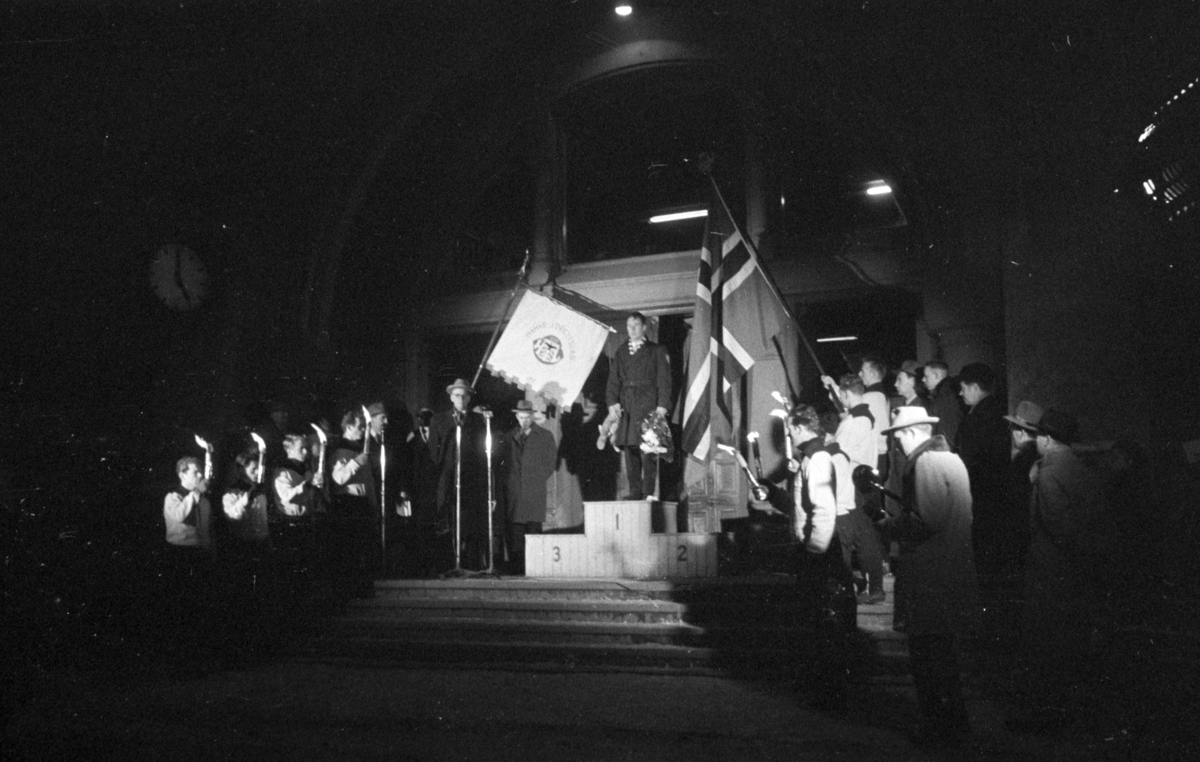 Egil Danielsen kommer hjem til Hamar etter å ha vunnet Olympisk gullmedalje i Spyd i Melbourne 1956. Hamar Jernbanestasjon. Danielsen på toppen av "seierspallen". Pressefotografer, fane Hamar Idrettslag, norsk flagg og fakler. 