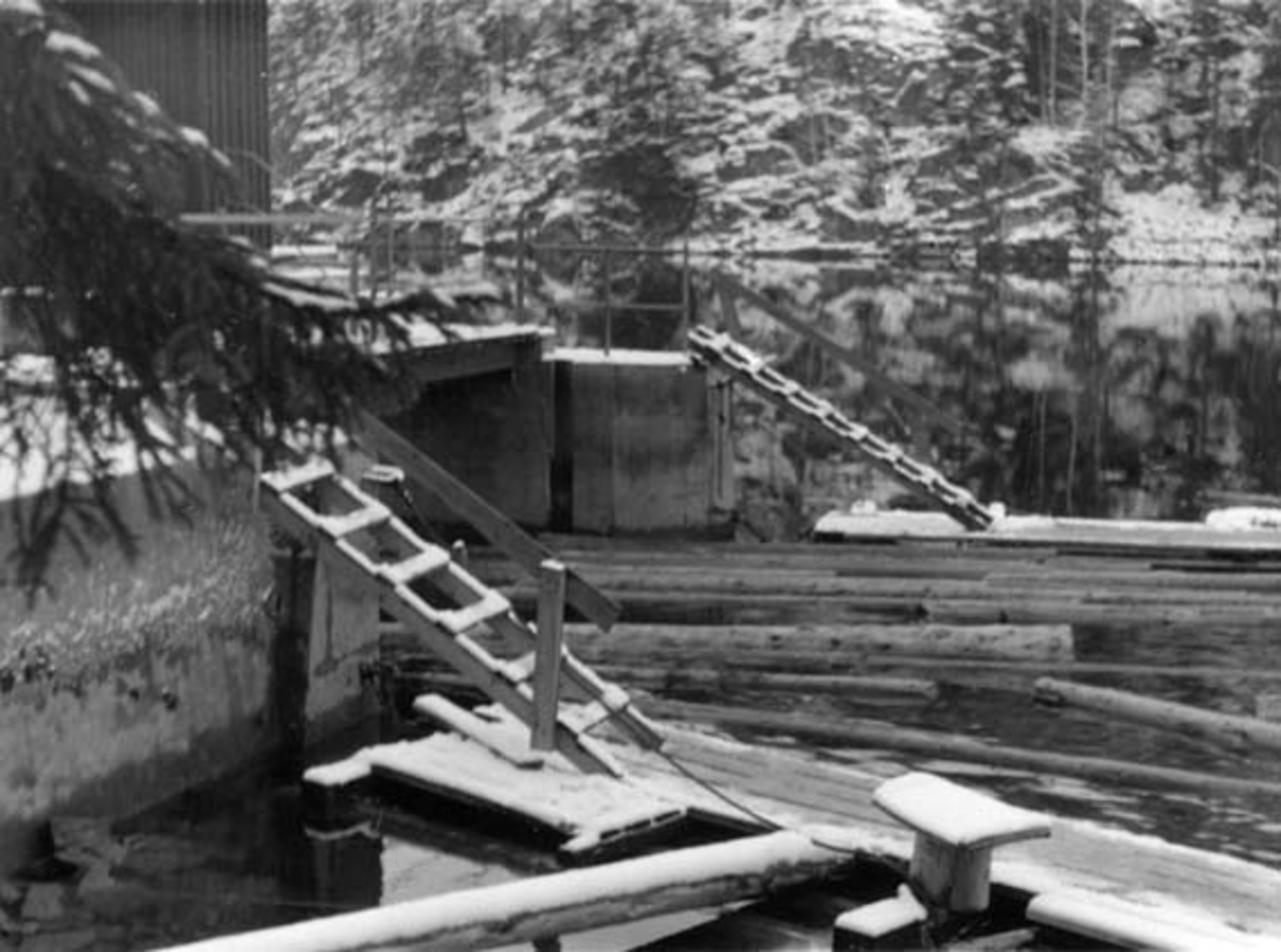 Inntaket til tømmerrenna ved kraftverket Gravfoss på Modum i Buskerud. Fotografiet viser en støpt damkonstruksjon med en bordkledd bygning på.  Bakenfor dammen ses et blankt vannspeil og ei strand med delvis snødekte bergskrenter og furuskog.  I forgrunnen ser en hvordan det er lagt flåter på begge sider av tømmerløpet.  Adkomsten til flåtene skjer via stiger, som er reist opp mot damkrona.  Fotografiet ble tatt i 1952. 