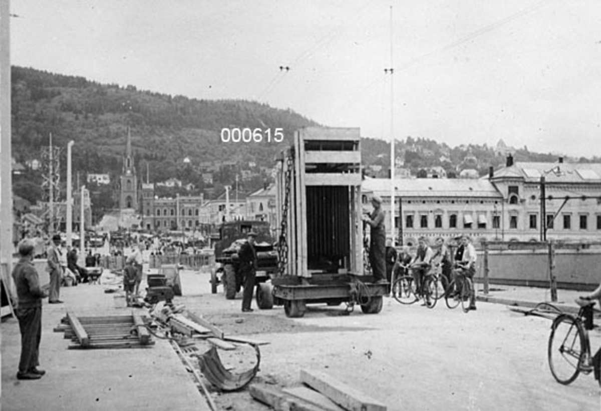 Transport av transformator som skulle installeres på A/S Buskerud Papirfabrikk på Åssida i Drammen (den gang i Lier kommune) i 1936.  Fotografiet er tatt på det som den gangen var ei ny bru over Drammenselva.  Transformatoren skulle åpenbart fraktes fra jernbanestasjonen over elva og videre på vegen mot Krokstadelva til en nådde fabrikken.  Ifølge et avisoppslag (jfr. SJF-F. 000618) var dette en transport som først ble mulig da det kom ny bru, fordi den gamle ikke var konstruert for å bære slike tunge lass.  Om denne transformatoren heter det for øvrig at den var tysk og veide 6 tonn. 

Fotografiet er tatt på et nytt brulegeme, der det ennå ligger en del trematerialer, muligens fra anleggsperioden, langs fortauskantene.  På fortauene står det en del menn og unggutter - de siste med sykler - og betrakter transporten, som skjer ved hjelp av en lastebil med etterhengt tilhenger, hvor transformatøren later til å være innebygd i ei kasseramme.  På den andre sida av brua ses forskjellige bygårder og kirka ved Bragernes torg, og bakenfor dette igjen hever en skogkledd ås (Bragernesåsen) seg over byen. 