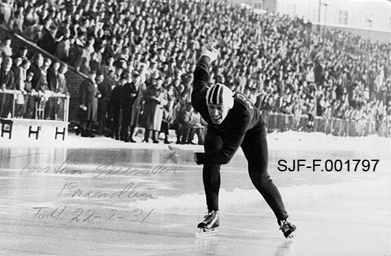 Skøyteløperen Torstein Seiersten (1931-2023) fra Brandbu på Hadeland, fotografert under et stevne på Bislett stadion i Oslo, antakelig i 1950-åra.  Seiersten er iført mørk, tettsittende trikot med «NORGE» med kvite bokstaver på ryggen.  På hodet har han ei tettsittende, kvit skøytelue med flaggfargene - rødt, kvitt og blått - i striper på fronten.  På hendene har han kvite strikkevotter.  Løperen har et innbitt uttrykk i ansiktet, og han veiver med armene, antakelig for å drive opp farten mot slutten av et løp.  I bakgrunnen står publikum tett på tribuner bak et nettinggjerde.  Over den lyse isflata i forgrunnen til venstre har noen skrevet «Torstein Seiersten, Brandbu, Født 22-2-31» med grønn kulepenn.
