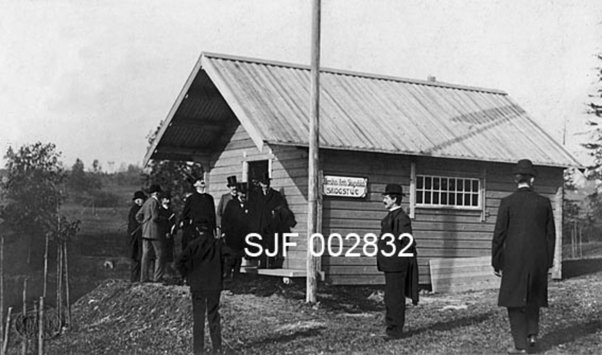 Eksteriøropptak fra den store jubileumsutstillingen på Frogner i Kristiania (Oslo) sommeren 1914.  Fotografiet viser ei tømmerkoie som er oppsatt av lafteplank.  Koia har inngangsdør i gavlenden og et smårutete, rektangulært vindu på den langveggen som vendte mot fotografen.  Saltaket er tekket med bord.  På veggen er det montert ei plate som forteller at det var Akershus Skogselskap som sto bak denne montasjen.  På kartongen museets kopi av fotografiet er montert på har noen skrevet "Høibraatens Tømmerkoie" med blyant.  Foran bygningen er det reist en stolpe som vi ikke ser toppen på, muligens ei flaggstang.  Da dette opptaket ble gjort hadde koia fornemt besøk.  Foran inngangspartiet ser vi nemlig ei forsamling av herrer med flosshatter, og midt blant dem står kong Haakon VII.  Det er grunn til å anta at bildet er fra en omvisning med kongen og sentrale menn fra utstillingskomiteen, kanskje også fra den øvrige nasjonale makteliten. 