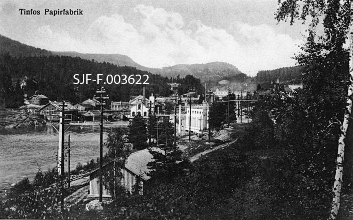 Industrianlegg nedenfor Tinnfossen i Notodden kommune i Telemark.  Bildet er fra et fotografisk postkort, som antakelig er fra omkring 1920.  Fotografen har stått i skråningen mot Tinnåvassdraget.  I forgrunnen vokste det krattskog med blant annet bjørk.  Litt nedenfor opptaksstedet lå det et enetasjes skur i bordkledd bindingsverk, med en veg eller skinnegang i forkant.  Industribebyggelsen, som overfor publikum skulle være motivet på dette kortet, befinner seg noe lengre inne i bildeflata.  Bebyggelsen vest for vassdraget (i venstre halvdel av bildeflata) tilhørte Tinfos Papirfabrik.  Her ser vi bygninger som er utført både i tre og i teglsteinsmur.  Teglbygningen med gavl rett imot fotografen er den første kraftstasjonen ved denne fossen, Tinfos 1 (1899-1900).  Over denne gavlen ser vi ei forholdsvis høy industripipe og gavlen på en større teglsteinsbygning, antakelig papirfabrikken.  Til høyre, og noe nærmere fotografen, ser vi en stilfull teglsteinsbygning med karakteristiske hjørnetårn og skyteskåraktig gesimsløsning.  Dette er kraftstasjonen Tinfos II.  I bakgrunnen skimtes skogkledde åsrygger. 