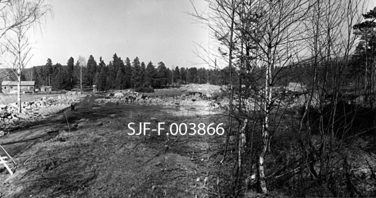 Fra opparbeidinga av tomteareal for Geithus Bruk på Modum i Buskerud. Dette bildet er sammensatt av to opptak, som begge er gjort i begynnelsen av april 1965. Da hadde man begynt å planere det arealet hvor industrihallen skulle reises. En del stein er samlet langs arealets ytterkant. Til venstre i bildet ligger et bolighus. Miljøet er ellers preget av skog.  Geithus Bruk var en virksomhet som kjøpte tømmer for barking og flishogging. Flisa ble levert til Skotselv Cellulosefabrikk i Øvre Eiker.