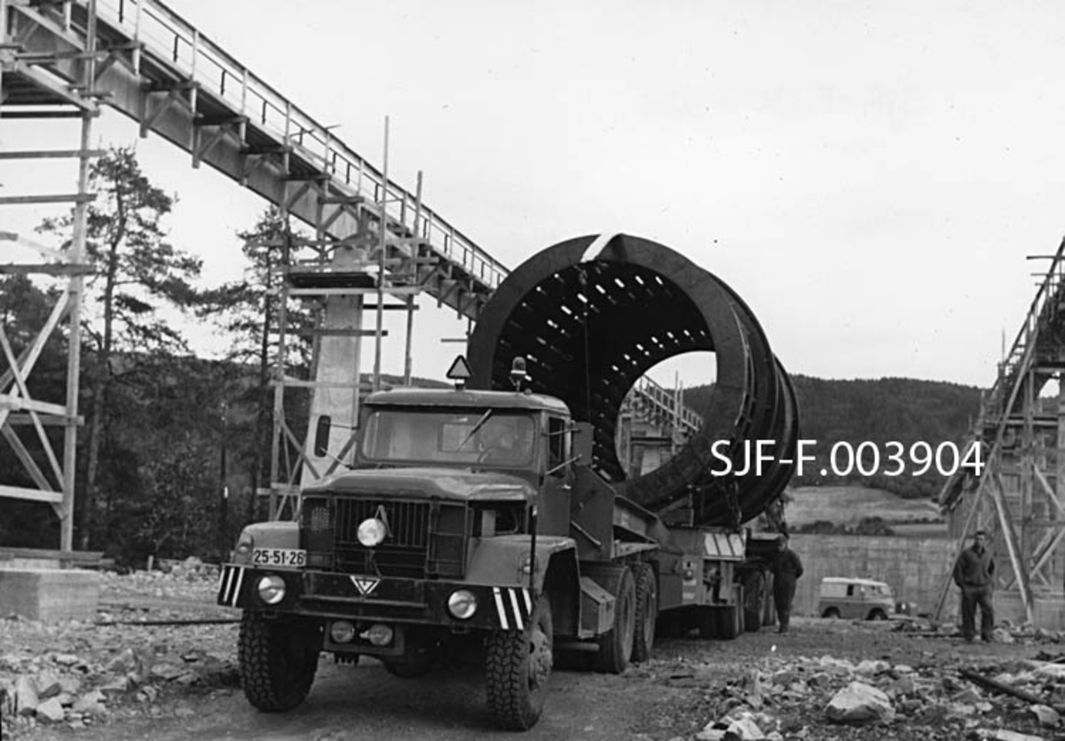 Transport av barketrommel til Geithus Bruk på Modum i Buskerud i 1966. Trommelen ble produsert ved A/S Myrens Verksted i Oslo.  På grunn av jernbaneunderganger og akseltrykkrestriksjoner kunne ikke trommelen transporteres hele vegen med lastebil.  Den ble derfor delt i to deler, hver med cirka 11 meters lengde, som ble transportert til Drammen med lastebåt.  Derfra ble det brukt lastebil, delvis om natta for å unngå å skape hindringer for annen trafikk.  Dette fotografiet er tatt i dagslys på et tidspunkt da lastebilen med en halv barketrommel sto parkert på det nye industriområdet, midt i den såkalte kranbanen.  Banen besto av to parallelle søylerekker med kraftige betongdragere på toppen, og den skulle brukes til å transportere tømmer fra Bergsjø i Drammenselva og fra lastebiler og jernbanevogner mot fabrikkbygningen.  Da dette fotografiet ble tatt var deler av forskalinga på kranbanen fortsatt intakt.  Det sentrale motivet i dette fotografiet er imidlertid den kraftige lastebilen med en 11 meter lang barketrommel med diameter på 3, 5-4 meter på lasteplanet.  På dette fotografiet aner vi ribbene i trommelen, som sammen med stokkenes gnissing mot hverandre i et fuktig miljø, skulle bidra til at barken ble revet av stokkene.  Da dette fotografiet ble tatt befant to menn seg på høyre side av lastebilen.  I bakgrunnen skimtes en åsrygg med dyrket mark nederst i liene og skog ovenfor. 