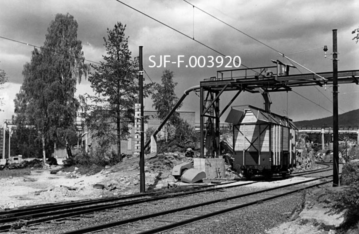 Lastestasjon for celluloseflis ved Geithus Bruk på Modum i Buskerud.  Fotografiet er tatt i 1967, da produksjonen ved denne bedriften startet. I forgrunnen ser vi det dobbeltsporete sidesporet mellom fabrikken og Geithus stasjon på Randsfjordbanen.  Ei flisvogn står under stålstativet på lastestasjonen, hvor flisa kommer i et rør fra fabrikken til venstre i bakgrunnen. Bakenfor lastestasjonen, til høyre i bildet, ser vi også noe av kranbanen, som ble brukt til å hente tømmer fra Bergsjø i Drammensvassdraget, fra jernbanevogner og lastebiler til fabrikken. Etter at tømmeret var kappet til kubb, barket og hogd til flis, ble sluttproduktet kastet over i en silo.  Derfra ble den transportert ned i en slusemater hvor flisa ble tatt av en luftstrøm som med en hastighet av 40 meter i sekundet blåste den mot lastestasjonen for jernbanevogner, som vi ser på dette fotografiet.  Her munnet flisrøret i et pendelrør som kunne beveges i vognenes bredde- og lengderetning, slik at flisa ble jevnt fordelt. Lastinga ble for øvrig fjernstyrt fra sorteringsavdelingen inne i fabrikkhallen, hvor det var en monitor som gjorde det mulig å følge oppfyllinga av vognene.  NSB leverte spesialvogner med høye karmer for denne transporten.  Hver vogn kunne ta 66 kubikkmeter flis.  Geithus Bruk skulle kunne fylle 15 fulle vogner hver dag.  Laste- og losseanlegget skal ha vært det første i sitt slag i Europa.  Flisa gikk stort sett til Skotselv Cellulosefabrikk i Øvre Eiker. 