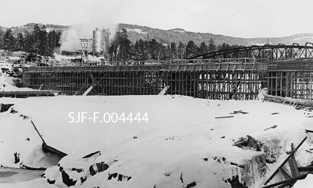 Bygging av ny dam på toppen av Geithusfossen på Modum i Buskerud.  Fotografiet er tatt 6. mars 1962.  Da var de bærende elementene, kraftige, støpte betongkar i elveløpet, på plass.  Det var også reist treforskaling for støping av dambrua.  Den nye dammen ble bygd vinteren etter at Union-konsernet hadde innviet Geithusfoss kraftverk.  Til dette anlegget ble det sprengt en inntakstunnel for vann fra et punkt ovenfor fossen, i fjellet under industriområdet til Drammenselvens Papirfabrikker.  Samtidig ble det også sprengt tømmerrenne utenom fossen.  I desember måned, et par måneder etter at kraftanlegget var ferdig, ble det lagt en fangdam i elvas hovedløp, nedenfor inntaket til kraftstasjonen og ovenfor fossen.  Ved å lede den beskjedne vintervannføringa gjennom de nye avløpene, kunne arbeidet med den nye dammen foregå på noenlunde tørr grunn.  Her er den nye dammen fotografert i motstrøms retning med et snødekt elveleie i forgrunnen.  Bakenfor skimter vi noe av buekonstruksjonen på jernbanebrua for sidesporet mellom Geithus stasjon på Randsfjordbanen og Drammenselvens Papirfabrikker. 