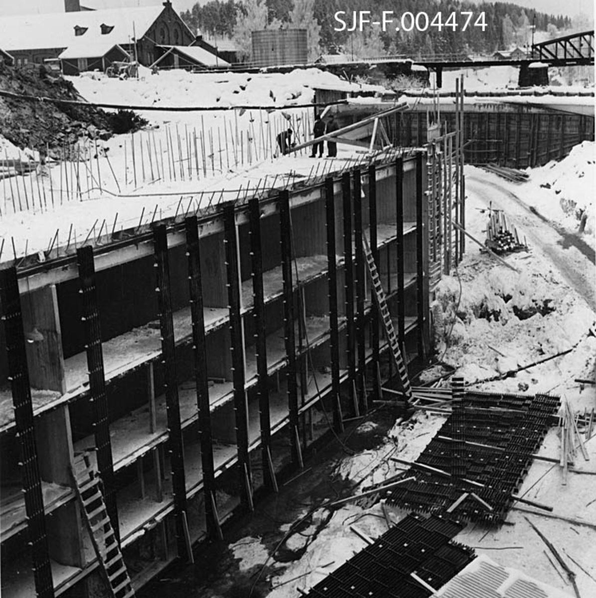 Fra vanninntaket til driftstunnelen for Geithusfoss kraftverk på Modum i Buskerud.  Fotografiet er tatt i februar 1961 og viser hvordan det var støpt et betongrammeverk med rektangulære åpninger ved det utsprengte innløpet (jfr. SJF-F. 004473).  På utsida av denne betongkonstruksjonen, mot vassdraget, var det montert kraftige stålskinner som skulle tjene som feste for varegrinder av samme materiale.  Da dette fotografiet ble tatt lå et par slike grinder på den snødekte bakken framfor vanninntaket.  Poenget var å sile vannet som kom inn i kraftverkstunnelen, slik at ikke tømmerstokker og andre objekter som kom flytende på elva fulgte med strømmen inn i driftstunnelen mot turbinen.  Fotografiet viser også hvordan det var støpt dekke over inntaksområdet.  Der skulle det legge skinner for ei kran (jfr. SJF-F. 004440) som skulle brukes til å betjene grindene og fjerne blant annet tømmerstokker, som ble liggende her.  I bakgrunnen til venstre ser vi et par av bygningene ved Drammenselvens Papirfabrikker, og til høyre litt av jernbanebrua på sidesporet mellom fabrikken og Geithus stasjon på Randsfjordbanen. 