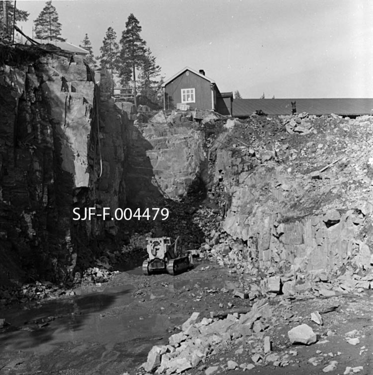 Inntaksområdet for driftstunnelen til Geithusfoss kraftverk på Modum i Buskerud, fotografert i anleggstida, nærmere bestemt i begynnelsen av august 1960.  Vi ser en shoveldoser i arbeid i et område der det er sprengt ut en stor dagåpning mot tunnelløpet, som gikk under spisebrakka ved Drammenselvens Papirfabrikker.  Vi der gavlen på denne bygningen, samt taket på det digre lagerskuret for ruller med avispapir og pakker med greaseproof (matpapir) oppå berget som det her arbeides med.  Gjennomslaget i tunnelen skjedde bare et par uker etter at dette fotografiet ble tatt.  Kraftstasjonen ble satt i drift 1. oktober 1961.  Da var inntaksområdet på dette fotografiet dekket av ei støpt betongsåle, og det var montert perforerte stålgrinder mot vassdraget (bak fotografen som har tatt dette bildet). 