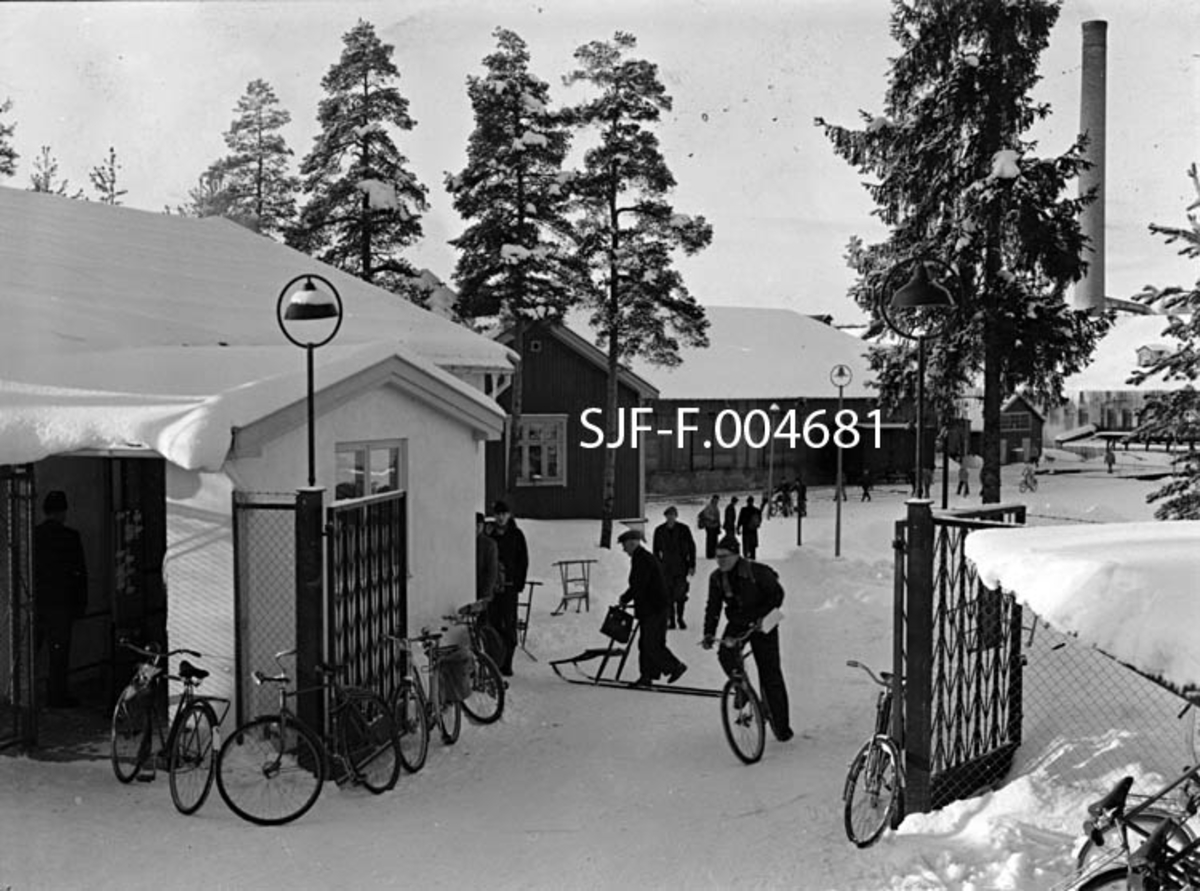 Fra porten til Drammenselvens Papirfabrikker på Modum i Buskerud.  Fotografiet er tatt ved arbeidstidas slutt en marsdag i 1957.  Vi ser en del av fabrikkens ansatte på vanding mot porten.  De fremste har nådd fram til sykler og sparkstøttinger som skulle forenkle hjemturen.  Til venstre i forgrunnen ligger den kvitmalte murbygningen fra 1954, som inneholdt bad og vasker for de ansatte og deres familier og kontor for portvakta.  Vi ser også at det var ei spesiell «sluse» foran portvaktrommet, der det hang et stemplingsur som mange av de ansatte måtte bortom på veg inn eller (som her) ut av fabrikken.  Ved den nevnte bygningen, like innenfor porten sto det en sykkel og fem sparkstøttinger.  Bak og til høyre for den kvite murbygningen ser vi gavlen på en panelt trebygning som lenge ble brukt som spisebrakke for fabrikkens ansatte, seinere som elektrokontor.  Bak denne igjen sto den store papirlagerbygningen med skyveporter mot jernbanesporet, som gikk langs langveggen mot innkjøringsvegen til fabrikken og elva.  Da dette fotografiet ble tatt sto ei av jernbanens godsvogner oppstilt foran lagerbygningen, antakelig klar for innlasting av papirruller.  Helt til høyre i bildet skimter vi en av fabrikkens teglsteinsmurte fabrikkhaller, samt den høye, sylindriske fabrikkpipa. 