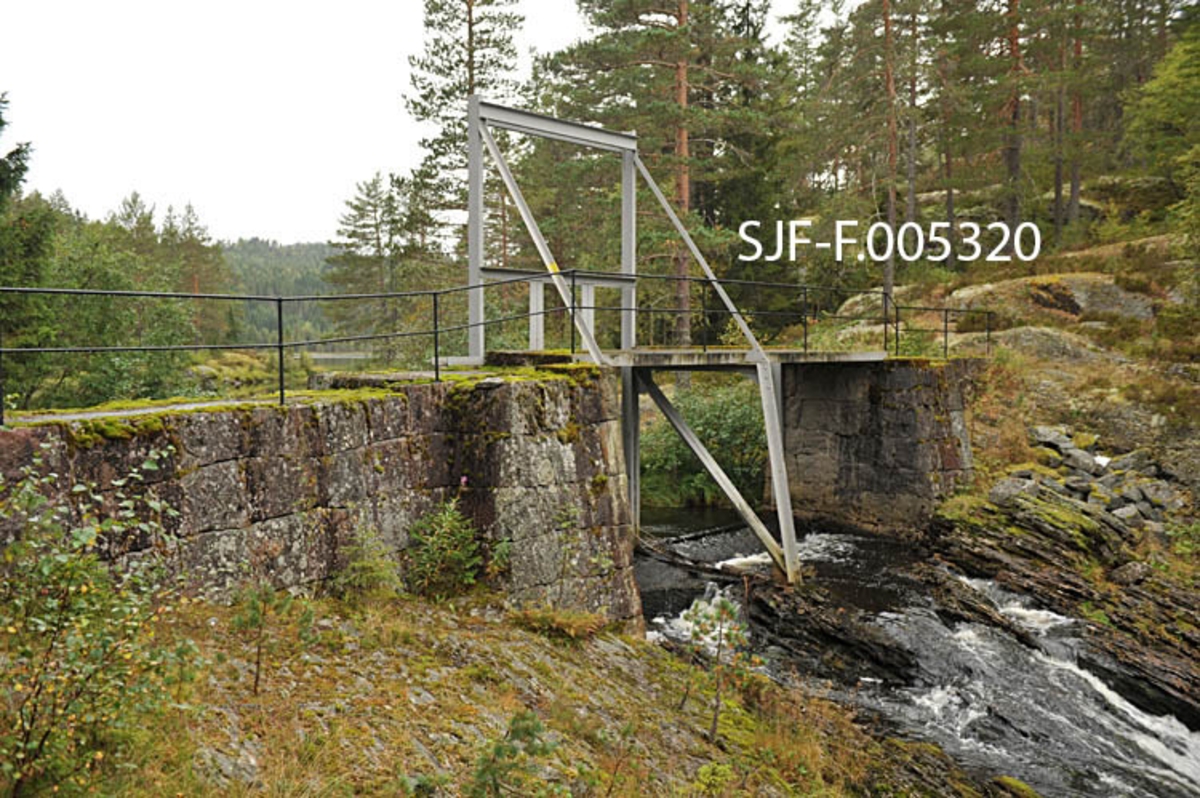 Vigdesjådammen ved utløpet av tjernet Vigdesjå i Kviteseid i Vest-Telemark.  Dammen er bygd av hoggen naturstein med krone av betong.  Dambrua har jernrekkverk på medstrøms side og ei stålramme for damlukene på motstrøms side.  Her ved utløpet av Vigdesjå renner Bygdaråi øst- og etter hvert nordøstover i bergrikt lende med Damåsan på nordsida og Kolakosi på sørsida, mot Høgfossen og Kolbjørnsrudfossen,  Fotografiet viser dammen omgitt av et landskap med blandingsskog (mye furu) og bergflater med lav- og lyngvegetasjon.  Bildet er tatt i vestlig retning mot damkonstruksjonen og damåpningen, hvor det er et visst fall i terrenget. 