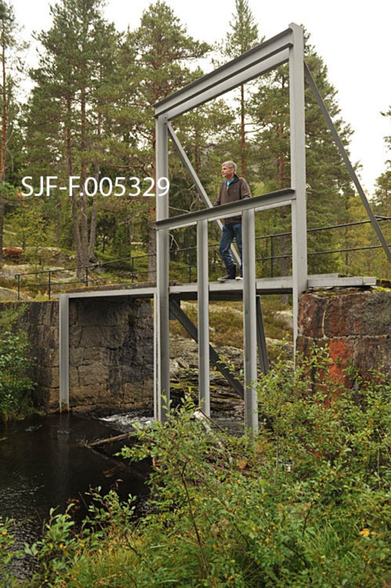 Vigdesjådammen ved utløpet av tjernet Vigdesjå i Kviteseid i Vest-Telemark.  Dammen er bygd av hoggen naturstein med krone av betong.  Dambrua har jernrekkverk på medstrøms side og ei stålramme for damlukene på motstrøms side.  Her ved utløpet av Vigdesjå renner Bygdaråi øst- og etter hvert nordøstover i bergrikt lende med Damåsan på nordsida og Kolakosi på sørsida, mot Høgfossen og Kolbjørnsrudfossen,  Fotografiet viser dammen omgitt av et landskap med blandingsskog (mye furu) og bergflater med lav- og lyngvegetasjon.  Bildet er tatt i nordlig retning, mot damkonstruksjonen og damåpningent Her var alle stengsler av tre borte på opptakstidspunktet, 40 år etter siste fløtingssesong.  Konservator Bjørn Bækkelund fra Norsk Skogmuseum sto på dambrua da bildet ble tatt. 