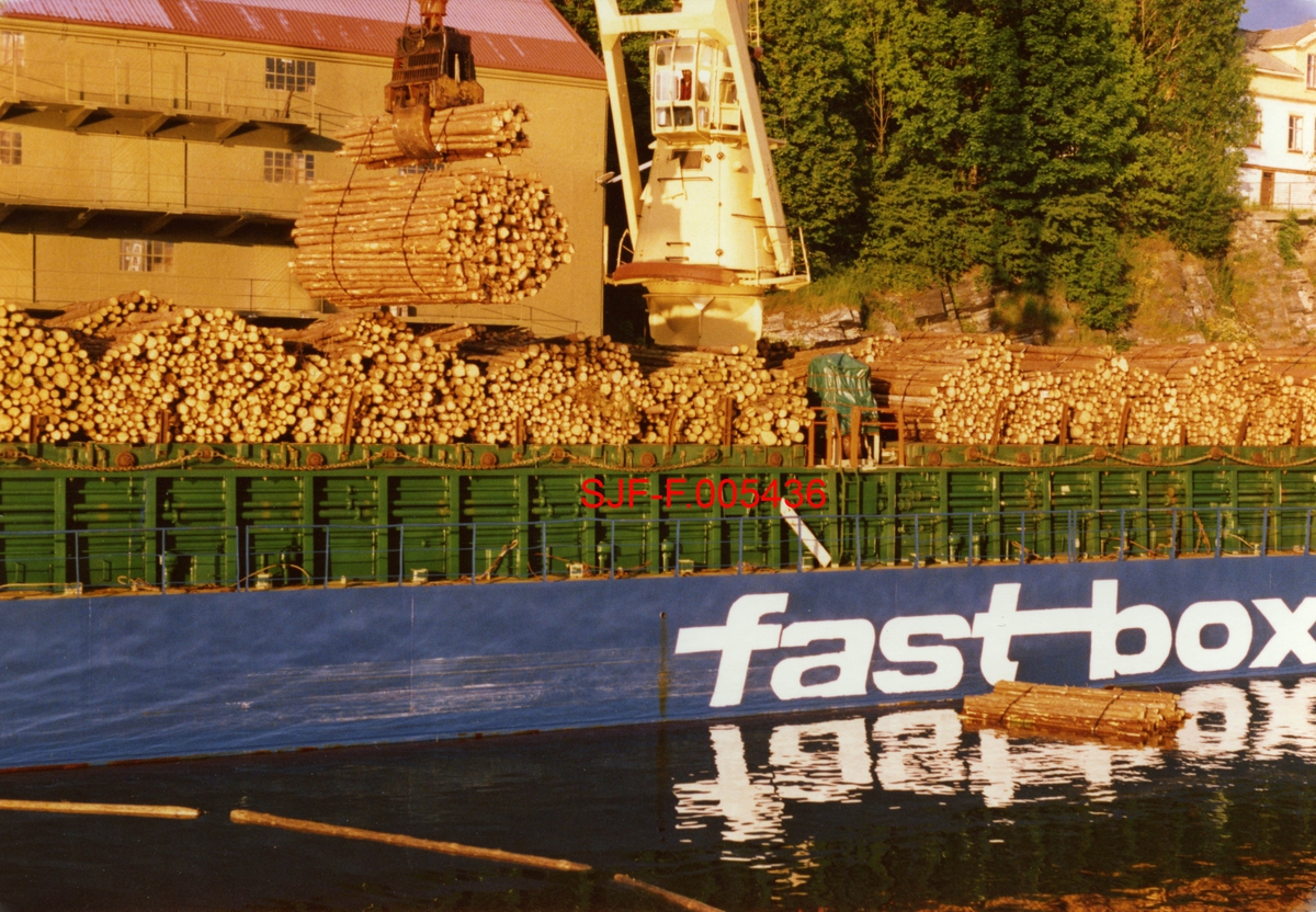 Lossing av tømmer fra fraktefartøy ved Bryggevannet, øverst i Skienselva.  Dette var tømmer som kom fra andre regioner.  Da dette fotografiet ble tatt (1986) kjøpte norsk treforedlingsindustri en god del råstoff fra Sverige.  Virket på dette fotografiet skulle brukes som råstoff for papirproduksjonen ved treforedlingsbedriften Union.  Fotografiet viser hvordan tømmeret ble løftet opp fra lektere og fraktebåter.  Deretter ble tømmerbuntene sluppet i Bryggevannet, i en «tekst» (ringbom).  Hver tekst kunne romme bortimot 100 bunter.  Når den den begynte å bli full av tømmer ble slept over til Unionfabrikken, på den andre sida av vassdraget og erstatte med en tom tekst.  Da dette fotografiet ble tatt hadde Skiensvassdragets Fellesfløtningsforening overtatt ansvaret også for mye av logistikken knyttet til transport av slikt virke inn på fabrikken.  På vannspeilet i forgrunnen ser vi (til venstre) en del av en «tekst» lense og (til høyre) en «bilmose», et sammenbundet tømmerlass et lastebilutislag ved Skiensvassdraget. 