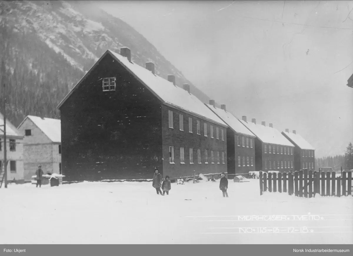 Fire murhus for flere familier stående på rekke på sørsiden av Tveito Alle. Barn stående i snøen i forgrunnen. I bakgrunnen pågår byggingen av de fire murhusene på nordsiden av Tveito Alle.