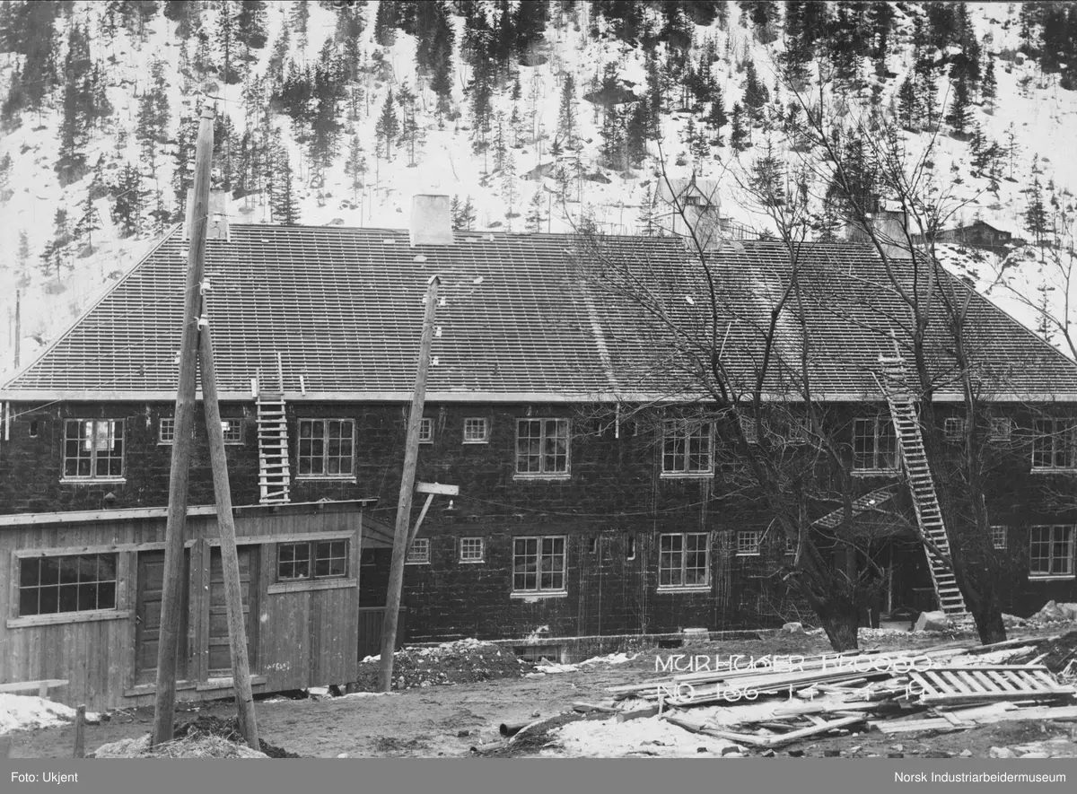 Murhusbebyggelsen på Krosso under oppføring. En bygning er ferdig reist med inngangsparti, dører og vinduer, samt fire piper. Taket har fått sløyfer og lekter, men ikke takstein. To stiger lent opp mot taket. Brakke i forgrunnen. Bolig på sørsiden av juvet langs Vemorksporet i bakgrunnen.