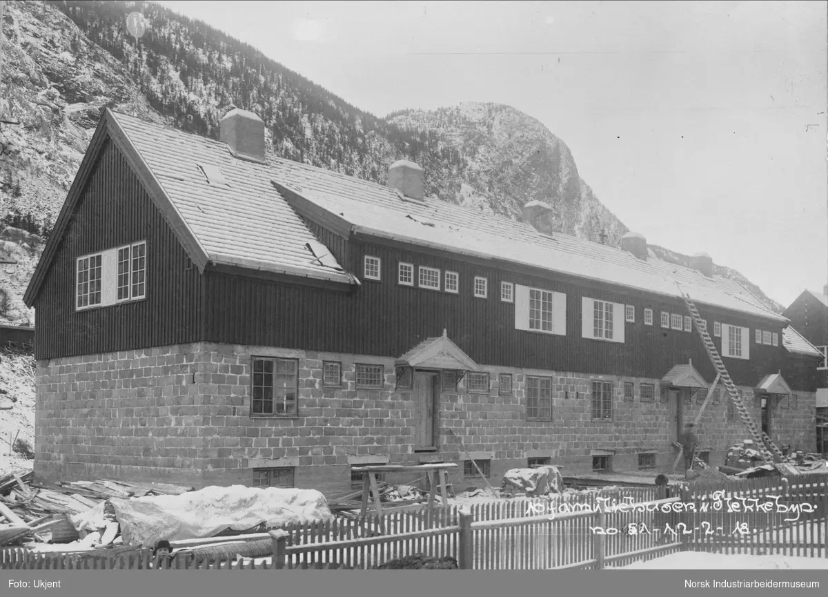 Mur og trehus i Flekkebyen for 10 familie bygges. Første etasje er bygget i mur, mens andre etasje er bygget i tre. Fasade mot Sam Eydes gate. Stiger står lent opptil husveggen. Fabrikkområdet i bakgrunnen.