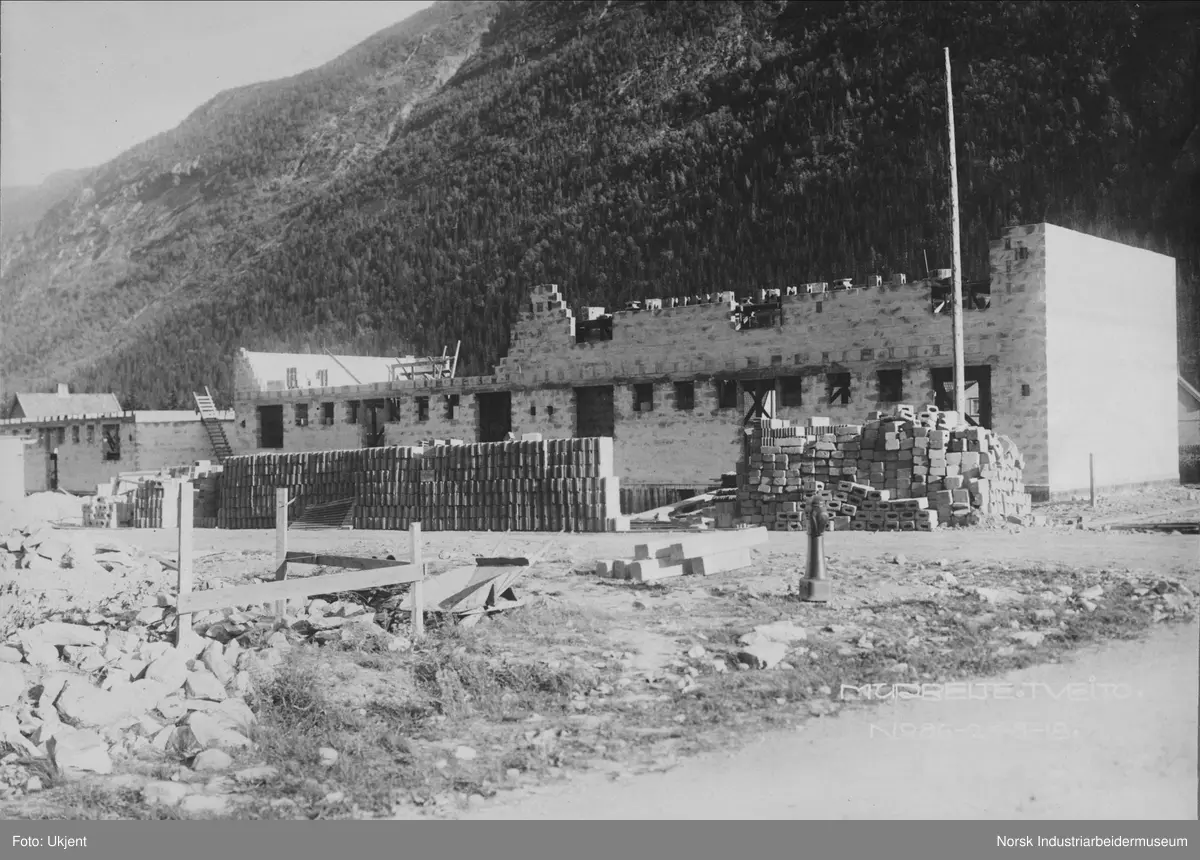 Murbebyggelse i Tveito Alle under bygging. På bolighusene på sørsiden av gaten bygges andre etasje. Brannhydrant stående på gatehjørne. Stabler av murstein stablet foran boligen.