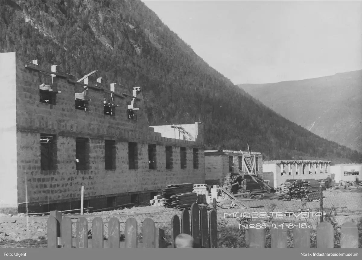 Bygging av murhus, flerfamiliehus, i Tveito Alle. Stabler med materialer og murstein liggende foran boligene. Gutt stående i port i forgrunn.