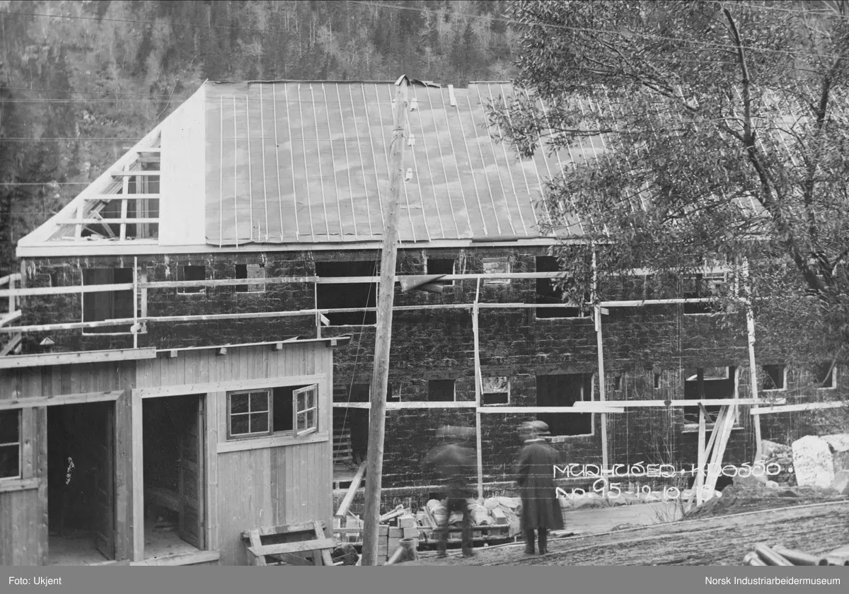 Murhusbebyggelsen på Krosso under oppføring.Murhusets første og andre etasje er reist. Arbeid med taket pågår, takbord, papp og sløyfer legges. To ingeniører ser på bygningen. Brakke ved siden av veien.