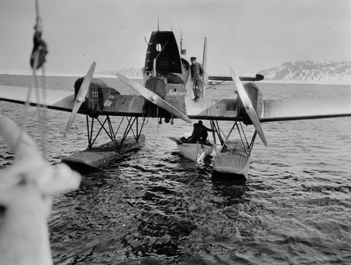 Sjöflygplan av Junkers-typ på vatten i fjällmiljö. En man på planet och en man i en roddbåt under.