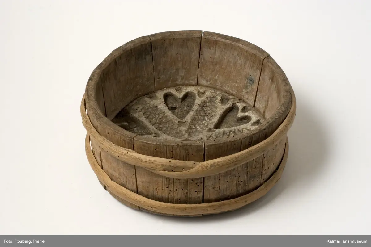 KLM 6627. Ostform. Av trä. Laggad, rund med två vidjeband, stavar av björk, botten av ek. Djupt skuren dekor i botten i form av grankottar och hjärtan i stjärnformation.