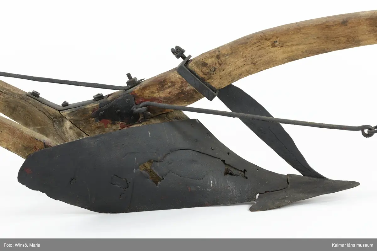 KLM 12824. Plog, trä, metall. Plog med handtag och ås av trä. Landsida av trä, järnskodd. Vändskiva och bill av järn. Tvärslå av järn mellan handtag och ås. Slåar mellan handtagen, två rundformade av järn, en U-formad av järn, en kantig av järn och en av trä. Längst fram på åsen en dragkrok av järn med reglage för inställning av höjd. På åsens mitt en fastskruvad rist. Risten är sammanfogad, eventuellt med färg, med billen. Tre långa krokar av järn hänger under åsen och möts i en järnring. Plogen är trasig på landsidan, träet är sprucket och järnbeslaget har lossnat. Plogen är rödmålad men färgen är kraftigt avskavd. Plogens järndetaljer är svartmålade med en röd eventuell rostskyddsfärg under. Den svarta färgen och rostskyddsfärgen är målad senare.