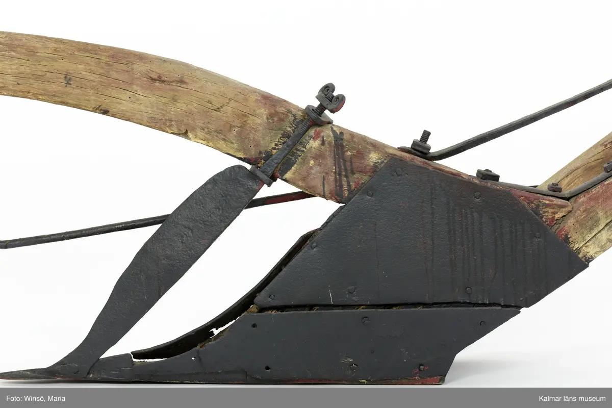 KLM 12824. Plog, trä, metall. Plog med handtag och ås av trä. Landsida av trä, järnskodd. Vändskiva och bill av järn. Tvärslå av järn mellan handtag och ås. Slåar mellan handtagen, två rundformade av järn, en U-formad av järn, en kantig av järn och en av trä. Längst fram på åsen en dragkrok av järn med reglage för inställning av höjd. På åsens mitt en fastskruvad rist. Risten är sammanfogad, eventuellt med färg, med billen. Tre långa krokar av järn hänger under åsen och möts i en järnring. Plogen är trasig på landsidan, träet är sprucket och järnbeslaget har lossnat. Plogen är rödmålad men färgen är kraftigt avskavd. Plogens järndetaljer är svartmålade med en röd eventuell rostskyddsfärg under. Den svarta färgen och rostskyddsfärgen är målad senare.