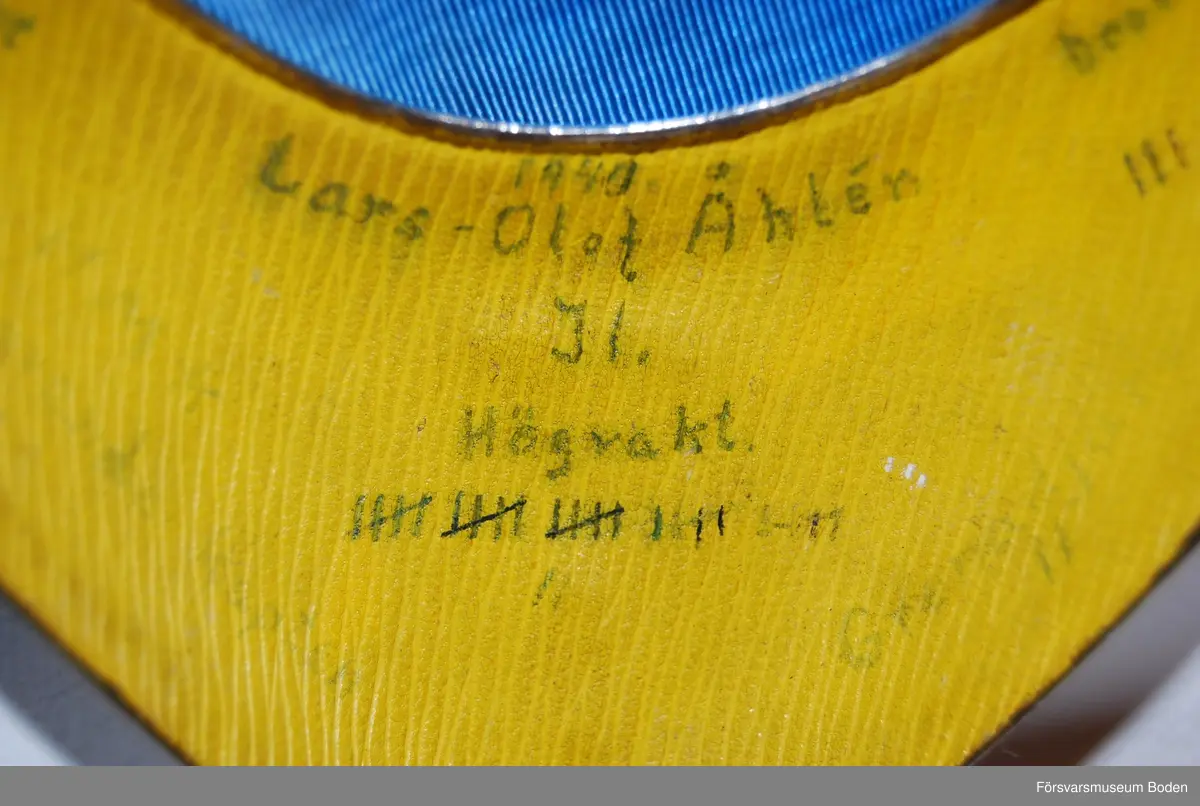 Av silver med blåemaljerad vapensköld och ljusblått band fäst med hakar. Baksidan fodrad med gult pappmaterial och med tidigare bärares påskrifter.