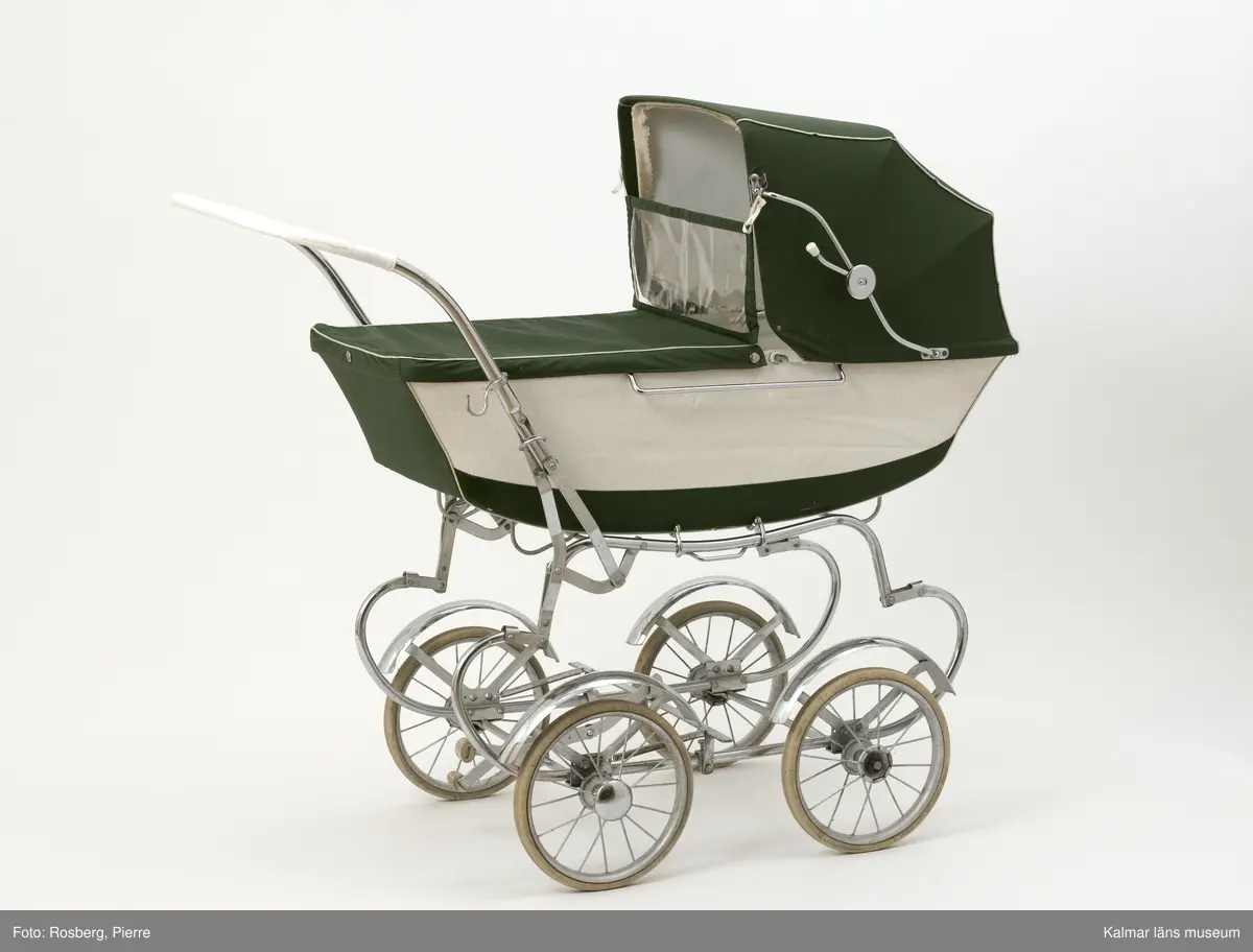 KLM 39807:1. Barnvagn, liggvagn. Tillverkad av Emmaljunga, Nr 119. Barnvagn med underrede av metall, gummiklädda hjul med text; Emmaljunga Trelleborg. Liggvagn med träbotten, sufflett och skyddstyg av grönt och vitt galonliknande material. Sömmarna är dekorerade med guldfärgad plast. Inuti vagnen en ligger en tillhörande madrass (KLM 39807:2).