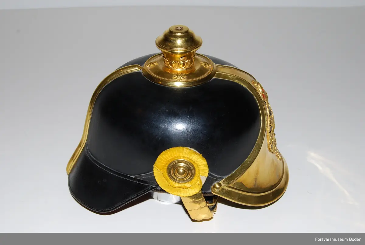 Svartlackerat läder med förgyllda beslag, framplåt, bakskena och hakrem. Förgyllt mössmärke med blå emalj. Gul sidenkokard under högra hakremsknappen. Med plym m/1885 av svart tagel, vilken användes vid stor paraddräkt. Pik medföljer ej.